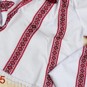 May include: White embroidered shirt with red and white geometric patterns. The shirt has detailed embroidery on the sleeves and bodice, with a lace trim at the hem. The number "No5" is visible in the bottom left corner.