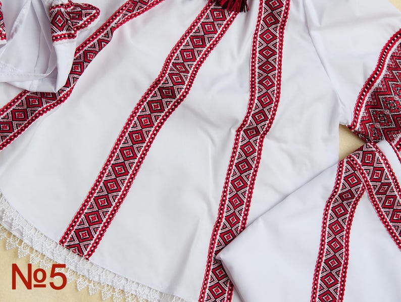 May include: White embroidered shirt with red and white geometric patterns. The shirt has detailed embroidery on the sleeves and bodice, with a lace trim at the hem. The number "No5" is visible in the bottom left corner.