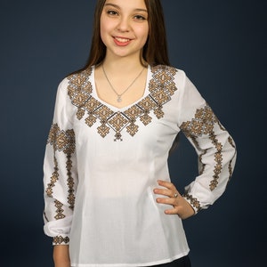 K&ouml;nnte beinhalten: Eine wei&szlig;e Bluse mit langen &Auml;rmeln und einem braunen Stickmuster. Die Bluse hat einen V-Ausschnitt und eine figurbetonte Silhouette.