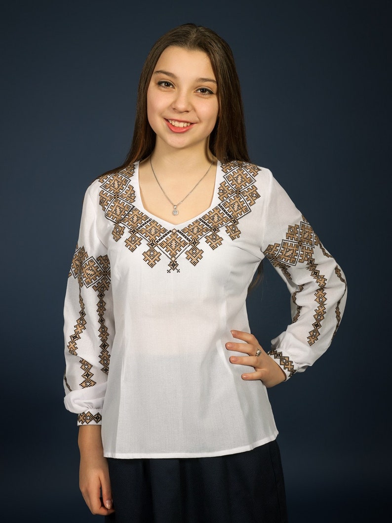 K&ouml;nnte beinhalten: Eine wei&szlig;e Bluse mit langen &Auml;rmeln und einem braunen Stickmuster. Die Bluse hat einen V-Ausschnitt und eine figurbetonte Silhouette.