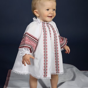 May include: A white baby dress with red and black embroidery on the sleeves and bodice. The dress features a lace trim and is worn by a young child. The background is a dark blue.