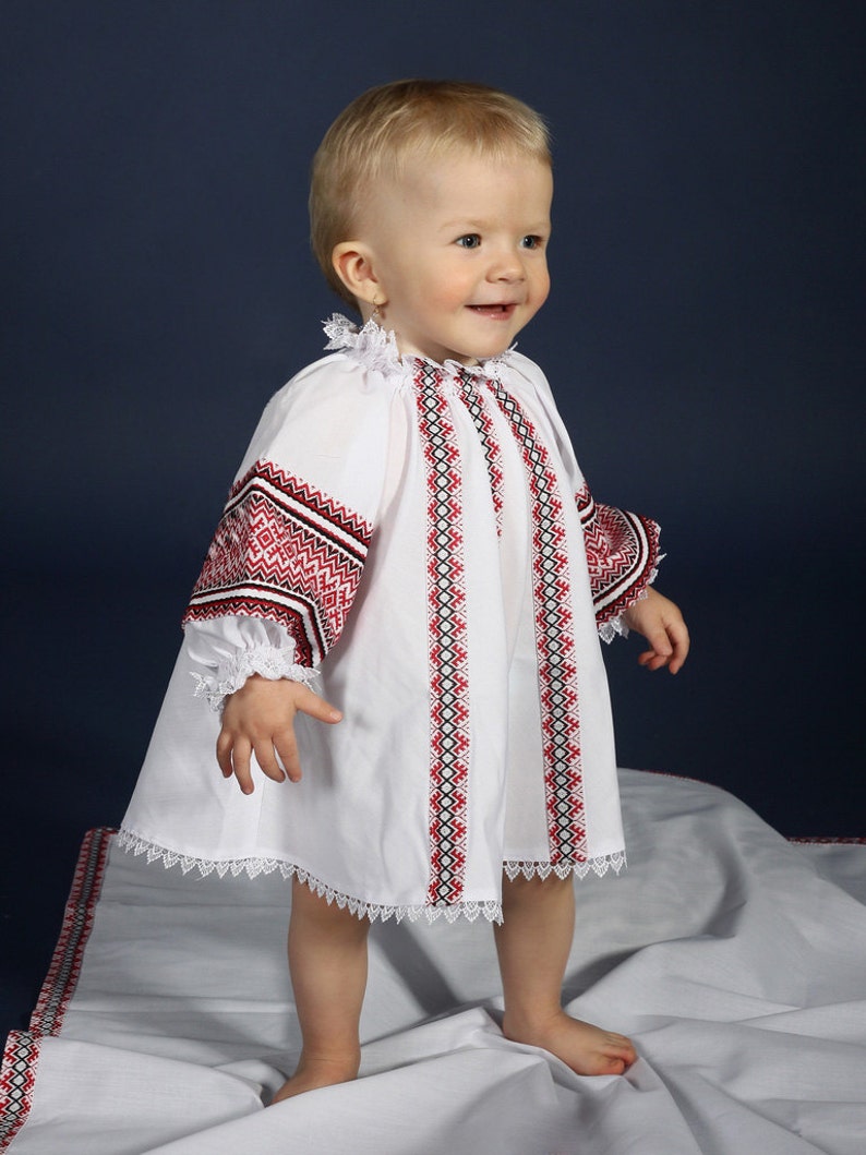 May include: A white baby dress with red and black embroidery on the sleeves and bodice. The dress features a lace trim and is worn by a young child. The background is a dark blue.