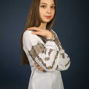 K&ouml;nnte beinhalten: Eine junge Frau mit langen braunen Haaren tr&auml;gt eine wei&szlig;e bestickte Bluse mit einem schwarzen und goldenen geometrischen Muster. Sie steht vor einem dunkelblauen Hintergrund.