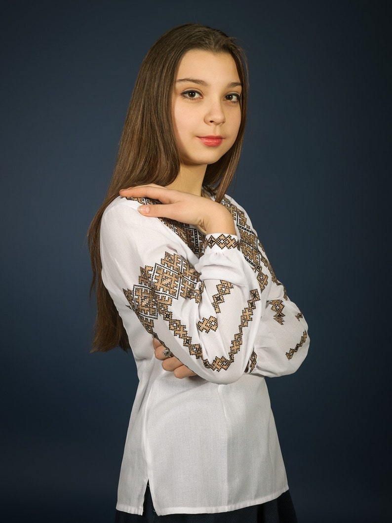 K&ouml;nnte beinhalten: Eine junge Frau mit langen braunen Haaren tr&auml;gt eine wei&szlig;e bestickte Bluse mit einem schwarzen und goldenen geometrischen Muster. Sie steht vor einem dunkelblauen Hintergrund.