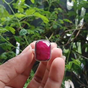May include: A silver ring featuring a large, rectangular, faceted deep pink gemstone. The stone is set in a polished silver bezel with a decorative twisted rope texture along the outer edge of the setting, creating a distinct border around the gem.