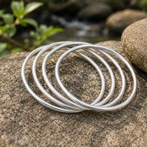 May include: A set of four thin, circular silver-colored bangles with a smooth, polished finish. The bracelets are stacked together, creating a layered appearance with a consistent metallic sheen across each individual band.