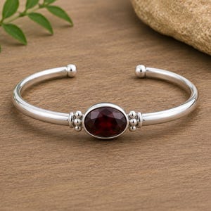 Op de afbeelding: Een gepolijste zilveren armband met een centrale ovaalvormige, gefacetteerde donkerrode edelsteen. De band is glad en afgerond en eindigt in kleine bolvormige uiteinden. Decoratieve clusters van kleine zilveren kraaltjes flankeren de zetting.