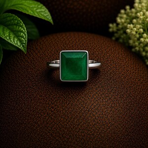 Peut inclure: Une bague argentée ornée d'une grande pierre précieuse vert foncé de taille carrée, sertie dans une monture en métal lisse. La bague possède un anneau fin et poli qui se connecte au sertissage carré, offrant un design symétrique et épuré.