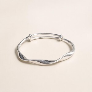 Può includere: Un bracciale rigido color argento caratterizzato da un design liscio e ondulato. Il bracciale ha una forma circolare con due piccoli terminali arrotondati dove si incontrano le estremità della fascia metallica, creando una struttura regolabile.