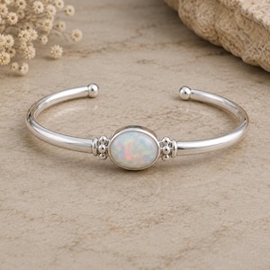 May include: A silver cuff bracelet featuring a central oval-shaped iridescent opal stone. The polished metal band has small spherical bead accents on either side of the stone setting and rounded terminals at the open ends of the bracelet.