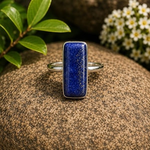 May include: A silver ring featuring a rectangular, deep blue lapis lazuli gemstone with visible gold-colored pyrite flecks. The stone is set in a smooth, polished silver bezel, mounted on a simple, slender silver band that encircles the finger.