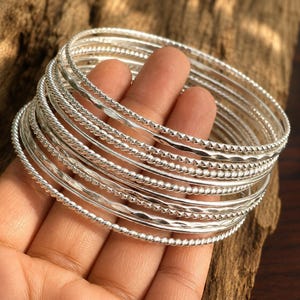 May include: A set of multiple silver-toned metal bangles featuring various textures, including smooth, twisted rope, and hammered finishes. These circular bracelets are stacked together, showcasing a reflective metallic surface and intricate detailing.