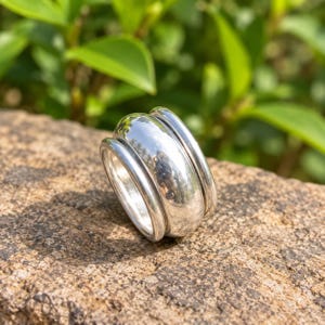 May include: A polished silver ring featuring a wide, smooth central band flanked by two thinner, rounded silver bands on either side. The metal surface is highly reflective, showing clear highlights and a smooth, rounded profile throughout the design.