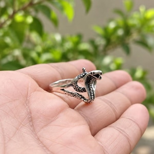 May include: A silver-toned metal ring featuring a detailed cobra snake design. The snake is coiled with its hood flared, showcasing textured scales and a defined head. The band of the ring is smooth and tapers towards the back, creating a continuous loop.