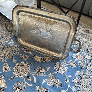 May include: A rectangular silver-toned serving tray featuring ornate, intricate floral engravings across the center and a decorative rope-style border. The tray includes two curved, detailed handles on opposite sides and exhibits a polished finish.