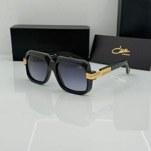 Könnte beinhalten: Schwarze Sonnenbrille mit texturiertem Schlangenleder-Effekt am Rahmen und verlaufenden Gläsern. Die Bügel haben goldfarbene Metallakzente und das Cazal-Logo ist auf dem Glas sichtbar. Präsentiert mit einem schwarzen Etui und einer Karte.