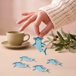 May include: A set of embroidered hammerhead shark patches in light blue and dark blue. Each patch features a smiling shark with a wide head and fins. The patches are flat, fabric-based designs intended for application onto various textile surfaces.