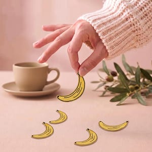 May include: A collection of small, yellow banana-shaped patches or pins. Each piece features a curved design with black outline details, mimicking the appearance of a peeled banana. The items are yellow with black accents.