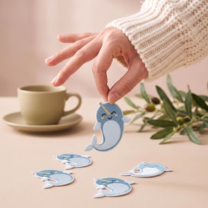May include: A set of embroidered narwhal patches featuring a light blue body, a white belly, and a gold-colored tusk. Each patch displays a smiling face with closed eyes and rosy cheeks, rendered in a simple, clean design for textile application.