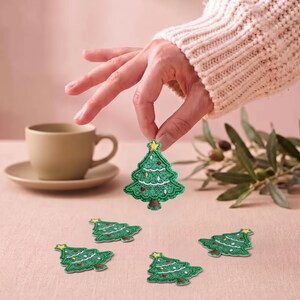 May include: A set of green Christmas tree patches featuring embroidered details, including a yellow star at the top, white garland accents, and small red and yellow ornaments. The patches have a brown trunk and a textured, stitched finish.