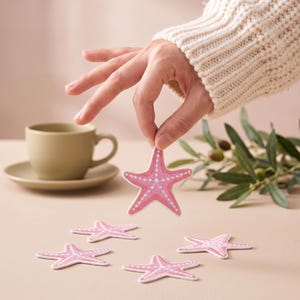 Peut inclure: Un ensemble de cinq écussons brodés en forme d'étoile de mer dans des tons rose et blanc. Chaque écusson présente une texture cousue détaillée avec un motif d'étoile centrale et des lignes rayonnantes. Les étoiles sont plates et fines.