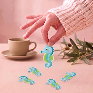 Op de afbeelding: Een verzameling geborduurde patches in de vorm van een zeepaardje met een kleurenpalet van lichtblauw en groen met witte accenten. Elke patch toont een zeepaardje in cartoonstijl met een gekrulde staart en een vin langs de rug.