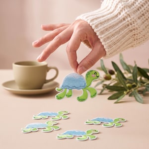 Pode incluir: Um emblema bordado de tartaruga com corpo verde-claro, carapaça azul-clara e um rosto sorridente. O emblema tem uma borda costurada e foi concebido como uma aplicação decorativa para tecidos, apresentando um design de tartaruga em desenho.