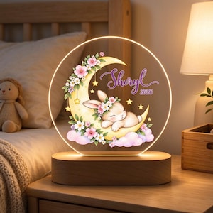 Puede incluir: Una luz nocturna circular de acrílico con base de madera que muestra un conejo durmiendo sobre una luna creciente amarilla, decorada con flores rosas y blancas, estrellas amarillas y nubes rosas. El texto dice Sheryl y el año 2026.