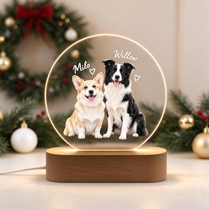 Puede incluir: Una luz nocturna circular de acrílico con la imagen impresa de un Corgi beige y un Border Collie blanco y negro. Los nombres Milo y Willow están impresos sobre cada perro con iconos de corazones. Se apoya en una base de madera.