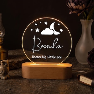 Könnte beinhalten: Ein kreisförmiges Acryl-Nachtlicht mit warmem gelbem Licht, das ein Mond-, Wolken- und Sternenmotiv über dem Namen Brenda und dem Text Dream big little one zeigt. Die klare Scheibe ist auf einem glatten, ovalen, hellen Holzsockel montiert.