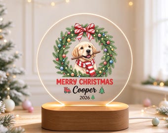 May include: A circular acrylic night light on a wooden base featuring a golden retriever puppy wearing a red and white striped scarf inside a Christmas wreath. The text reads Merry Christmas, Cooper, and 2026, with small icons of a red train and green trees.