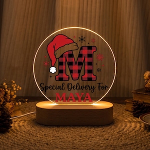 Può includere: Una luce notturna circolare in acrilico con base in legno che presenta una lettera M a quadri rossi e neri sormontata da un cappello da Babbo Natale. Il testo recita Special Delivery For MAYA in caratteri neri e rossi con fiocchi di neve.