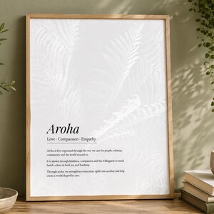 Può includere: Una stampa da parete rettangolare in una cornice di legno chiaro con sfondo bianco e un sottile motivo a foglie di felce pallide. Il testo recita: Aroha, Love, Compassion, Empathy, seguito da una definizione della parola Aroha in nero.