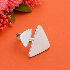 May include: A silver-toned ring featuring two distinct triangular elements of different sizes. The metal surface has a matte, brushed texture, and the two geometric shapes are connected by a thin band that wraps around the finger.