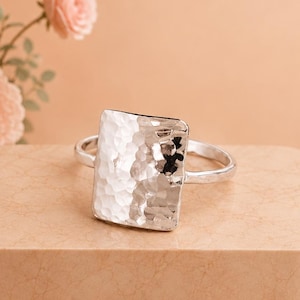 Könnte beinhalten: Ein silberner Ring mit einer großen, rechteckigen, gehämmerten Metalloberfläche, die eine reflektierende und strukturierte Textur aufweist. Die Ringschiene ist dünn und kreisförmig und passt zum Silberton des rechteckigen Mittelteils.