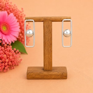 May include: A pair of silver-toned earrings, each featuring a vertical rectangular frame with a smooth, polished sphere suspended in the center. The earrings are displayed on a wooden T-shaped stand, showcasing a clean, geometric design.