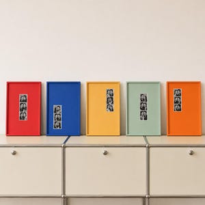May include: Five rectangular picture frames in red, blue, yellow, sage green, and orange, each featuring a vertical strip of three black and white photo booth portraits centered on a solid colored background within the frame.