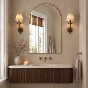 May include: An arched wall mirror with a thin gold frame hangs above a floating vanity featuring dark brown vertical slats. The vanity has a white countertop with a built-in sink, a gold faucet, two wall sconces, a towel ring, and a vase.