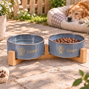 Peut inclure: Deux bols pour animaux de compagnie en céramique bleue, posés sur un support en bois clair. Chaque bol présente une empreinte de patte dorée avec un nœud et le nom Cooper inscrit en doré. Un bol contient de l'eau et l'autre des croquettes.