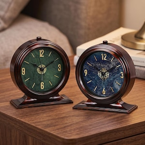 May include: Two round table clocks with dark brown frames and rectangular bases. One clock features a green marbled face, while the other has a blue marbled face. Both clocks display gold-colored numerals at 3, 6, 9, and 12 o'clock.