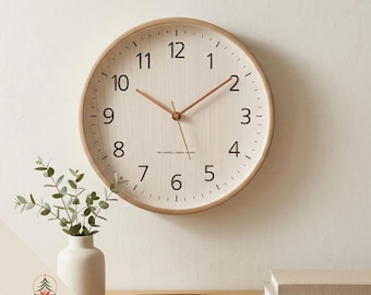 May include: A round wall clock with a light wood frame and a cream-colored face featuring black Arabic numerals from 1 to 12. The clock has thin copper-colored hour and minute hands and a thin second hand, with small text printed near the center.