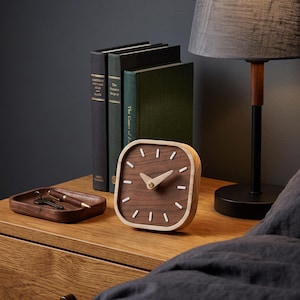 May include: A square wooden desk clock with a dark walnut face, light wood frame, and white hour markers. The clock features two light wood hands and a small brass center pin. Beside it sits a small wooden tray containing a metal key and a pen.