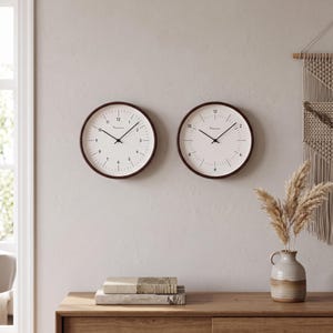 Puede incluir: Dos relojes de pared circulares con marcos de color marrón oscuro y esferas de color blanco roto. Cada reloj tiene manecillas negras para horas y minutos, números negros del 1 al 12 y marcas negras para los minutos. El texto "KASSEL" aparece impreso.