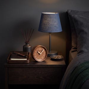 Op de afbeelding: Een ronde analoge houten klok met lichtgekleurde cijfers en wijzers, naast een tafellamp met een blauwe stoffen kap, een kleine donkere vaas met geurstokjes, een houten dienblad met een notitieboek en pen, en een klein schaaltje met ringen.