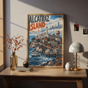 May include: A framed travel poster featuring a stylized illustration of Alcatraz Island with the text ALCATRAZ ISLAND in bold lettering. The artwork depicts the island, a lighthouse, a water tower, a ferry boat, and a distant city skyline.