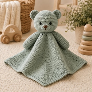 May include: A sage green crochet security blanket featuring a central bear head with dark eyes and a brown nose. The bear has two small rounded ears and a soft, textured body that flares out into a square-shaped blanket with a stitched border.
