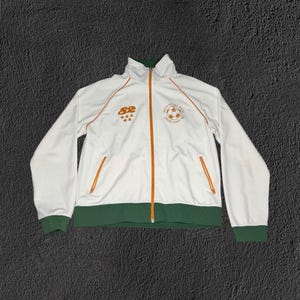 May include: A white zip-up track jacket with a dark green hem and cuffs. It features orange piping, an orange zipper, and orange graphics including the number 82, five stars, and a soccer ball logo with the text Golden Goal Rec Center Soccer League 1982.
