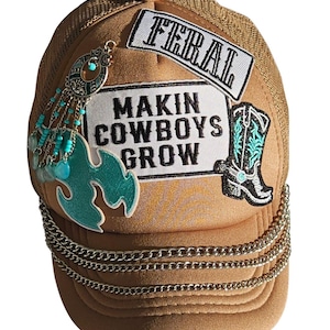 May include: A tan trucker cap with a mesh back and a front panel featuring patches reading FERAL and MAKIN COWBOYS GROW. The cap is decorated with a black and white cowboy boot patch, a teal flame-shaped patch, silver chains, and turquoise beads.