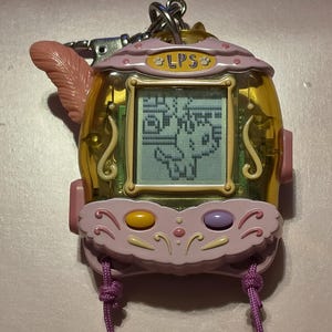 May include: A handheld electronic pet game with a translucent yellow casing and a light pink decorative frame. The device features a digital screen displaying a pixelated character, a metal keychain attachment, a pink feather accent, and two buttons.