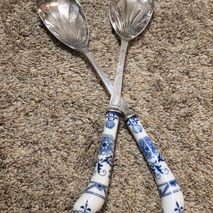 May include: A pair of silver-plated salad servers featuring polished metal bowls with fluted detailing and curved white ceramic handles decorated with intricate blue floral patterns and geometric bands in a classic, elegant design.